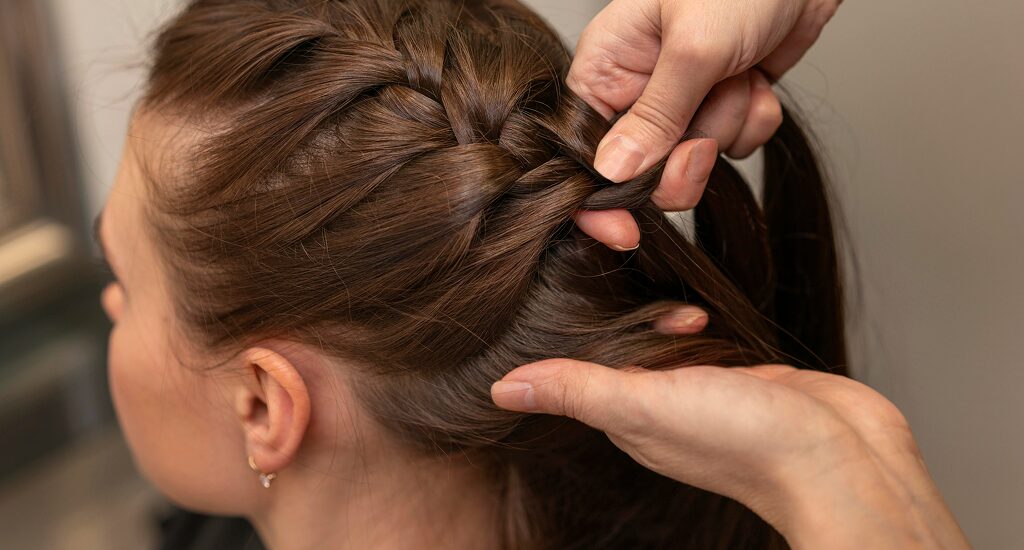 A close-up of a person's hands skillfully weaving a French braid into another person's long, dark blonde hair, showing the detailed cross-over technique.