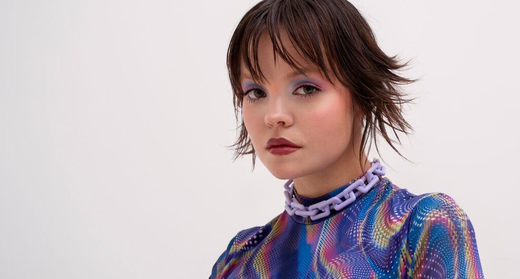 A young woman with a short, layered brown mullet and purple eyeshadow looks into the camera, wearing a vibrant mesh top and a chunky lilac chain necklace.