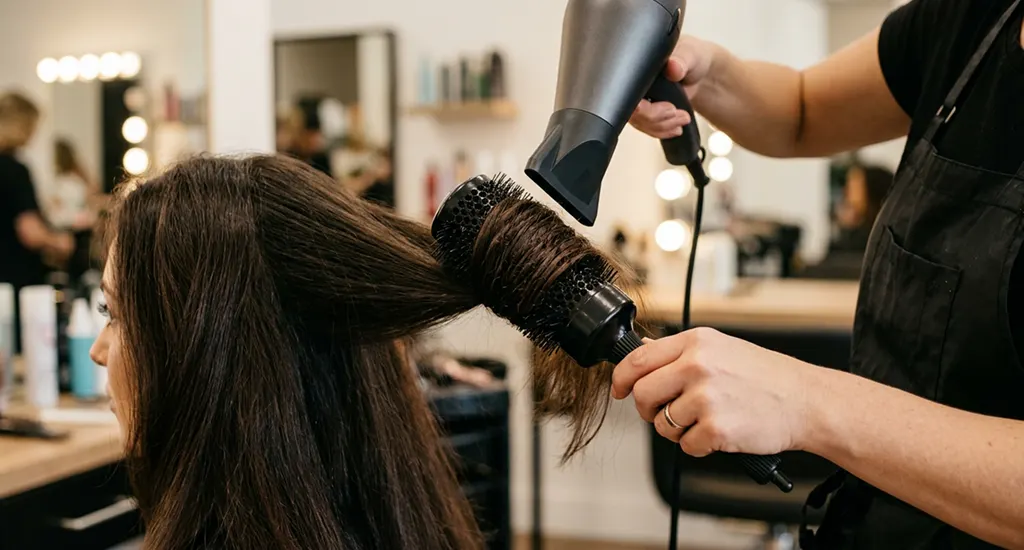 A professional stylist blow-drying a client's thick dark hair using a large round brush and a concentrator nozzle.