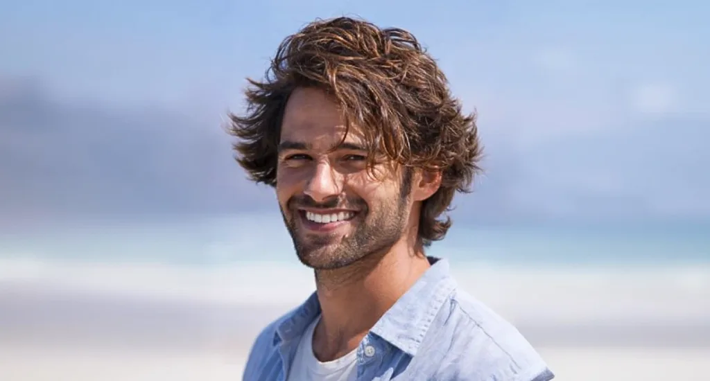 A smiling man outdoors near a beach backdrop showcasing a medium-length layered haircut with sun-kissed, natural brown waves.